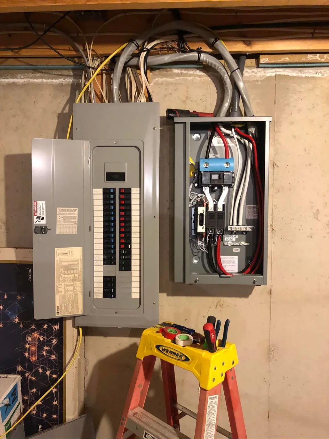 An electrical panel and subpanel with open covers showing circuit breakers and wiring are mounted on a basement wall. A red ladder with tools placed on top is positioned in front of them.